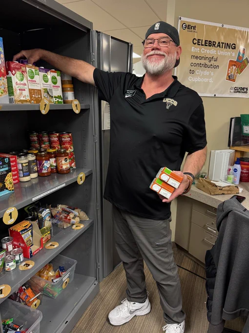A person is standing by an open pantry cabinet filled with various food items, holding some boxes and reaching for more. They are wearing a black UCCS shirt and a hat. A sign in the background reads, "Celebrating Ent Credit Union's meaningful contribution to Clyde's Cupboard.