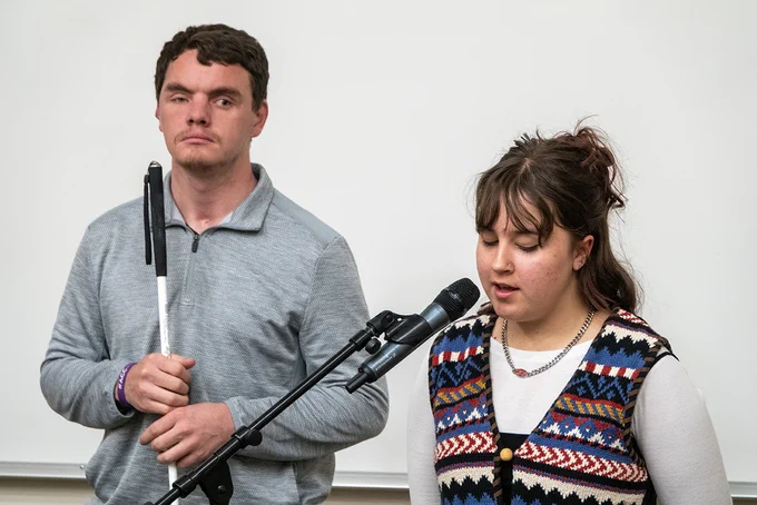 A man holding a white cane stands next to a woman speaking into a microphone against a plain background.