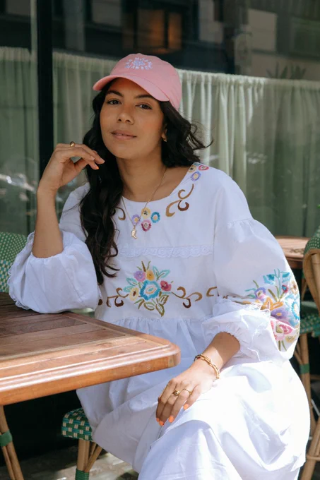 Annie Phillips wearing a white dress with colourful floral prints and a pink cap. She is sitting at a table, leaning on one elbow. 