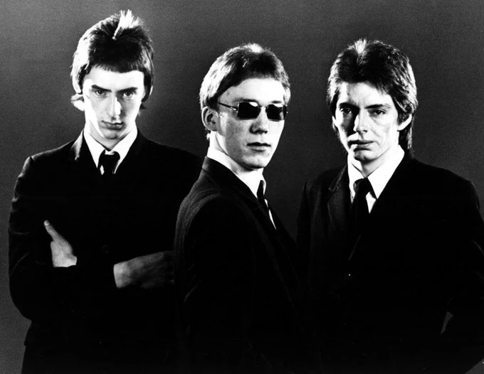 The Jam in a black and white promotional photo circa 1980, featuring Paul Weller (centre), Bruce Foxton (left), and Rick Buckler (right), dressed in sharp mod suits with a stark, minimalist backdrop reflecting their early post-punk era.