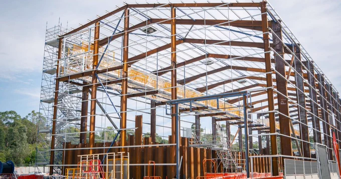 A large metal framework of a building under construction is shown. Scaffolding is visible on the left side. Trees and a cloudy sky form the background.