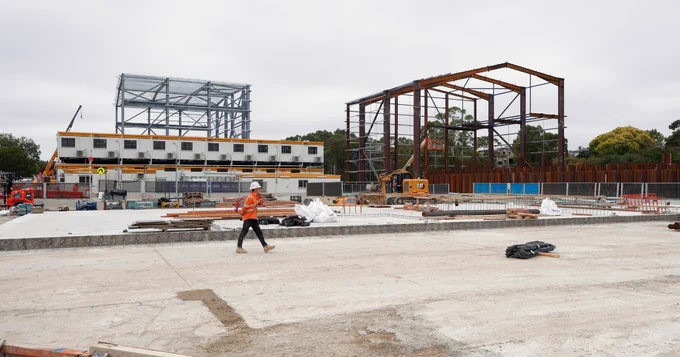 A construction site with two large steel frameworks and a row of portable buildings in the background. A person in a safety vest walks across the foreground. Various construction vehicles and materials are scattered around the site.