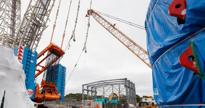 A construction site with a large crane lifting materials. In the foreground, machinery wrapped in blue tarps. A steel structure is partially built in the background, surrounded by various construction equipment and materials.