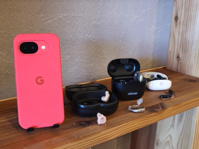 ピンクのケースに入ったGoogle Pixel 10aと、
Starkey Genesis AI、Oticon Intent、
オーダーメイドの耳あな型補聴器3台を
木の棚に並べた写真。