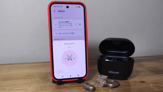 Google Pixel 10aの画面に補聴器のペアリング画面が
表示されている様子。隣にはOticonの充電ケースと
オーダーメイドの耳あな型補聴器が置かれています。
補聴器とスマホがGoogle Fast Pairで接続しようとしています。
