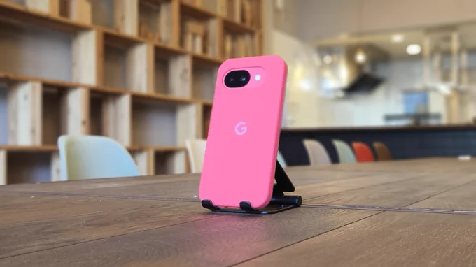 たかまつトコトコの店内でGoogle Pixel 10aを
スタンドに立てて撮影した写真。背面はピンクのケースで、
カメラ部分が完全フラットなデザインが見えます。
背景には木の棚と本、椅子が並んでいます。