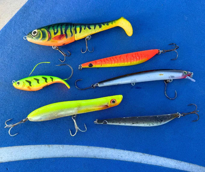 Six colorful fishing lures arranged on a blue surface, featuring various shapes and vibrant patterns.