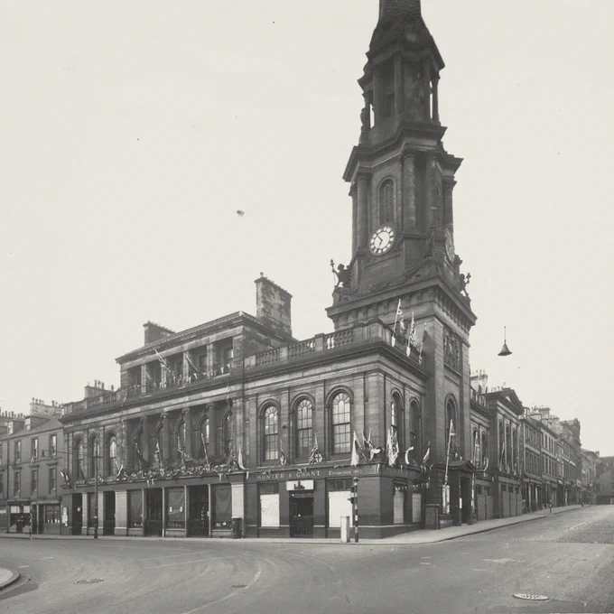 Black‑and‑white historic photograph of Ayr Town Hall seen from a street corner. The large stone building features arched windows, decorative façades, and a tall clock tower rising above the roofline. Surrounding streets appear quiet, with neighbouring buildings visible along the road.