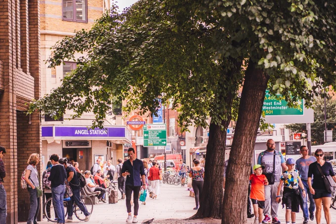 A street scene near Angel Station in London. People are walking along the sidewalk, with some sitting on benches. There are bicycles parked nearby, and a large tree provides shade. Various road signs are visible in the background.