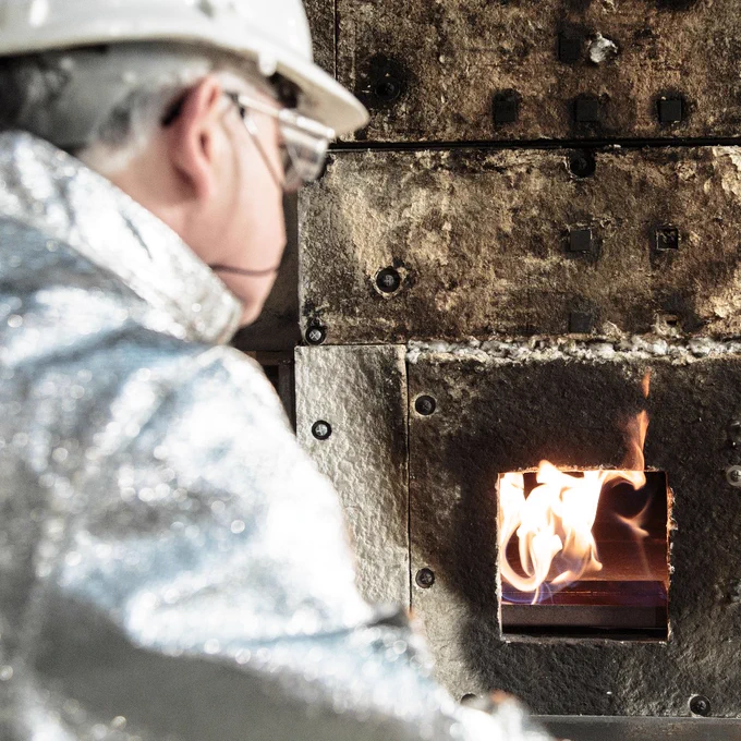 Worker in protective gear monitoring a small furnace flame inside an industrial kiln.