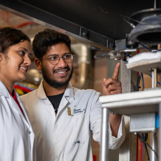 Two scientists in white lab coats examining a mechanical device in a laboratory setting with industrial equipment in the background.