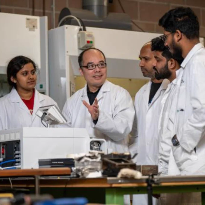 Four scientists in white lab coats discuss data around laboratory equipment in an industrial research setting.