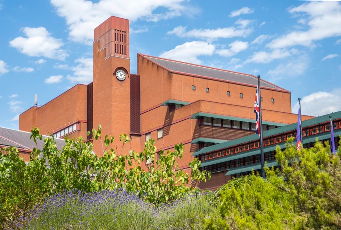 A red brick brutalist building with tall clock tower against a blue sky.