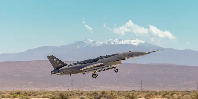 Military jet fighter taking off from desert runway with mountains in the background under clear blue sky.
