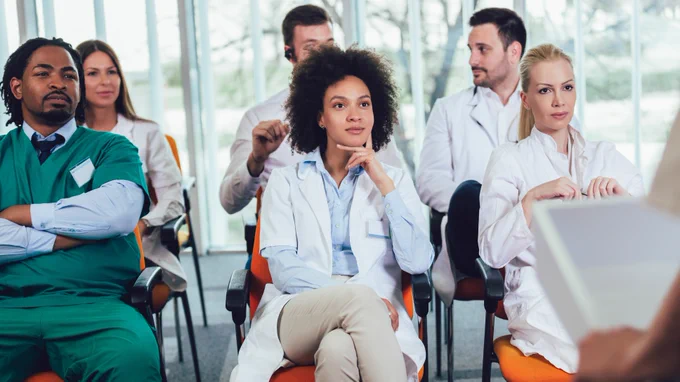 A group of medical professionals in uniforms is seated in a classroom setting, attentively listening to a speaker. 