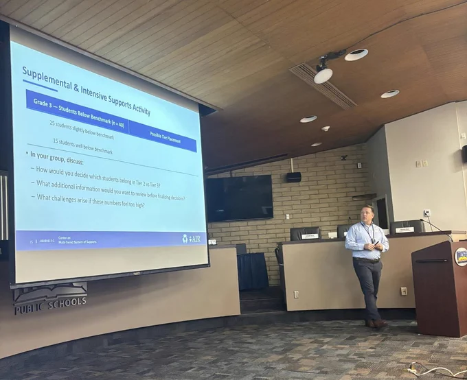 MTSS Center staff member Kyle Allen stands at a podium in a board room beside a projected slide titled “Supplemental & Intensive Supports Activity,” showing Grade 3 benchmark data and discussion prompts. The slide includes charts and bullet points, with the MTSS Center logo at the bottom. 