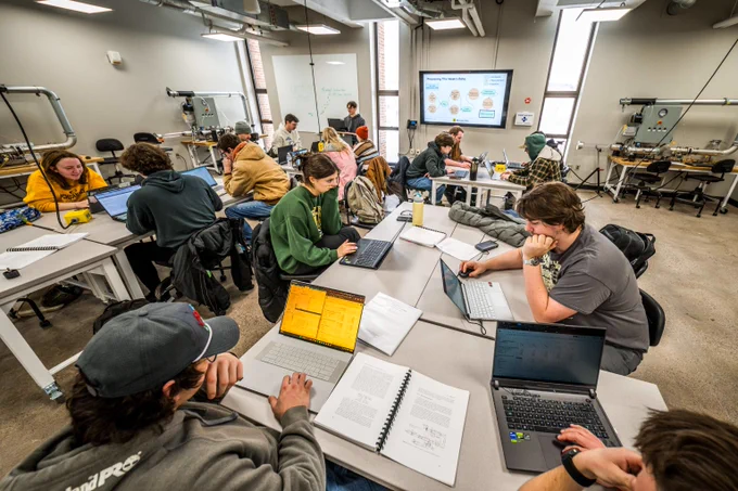 Students collaborate on laptops and notebooks in a bright, modern classroom with technical equipment and a presentation screen.