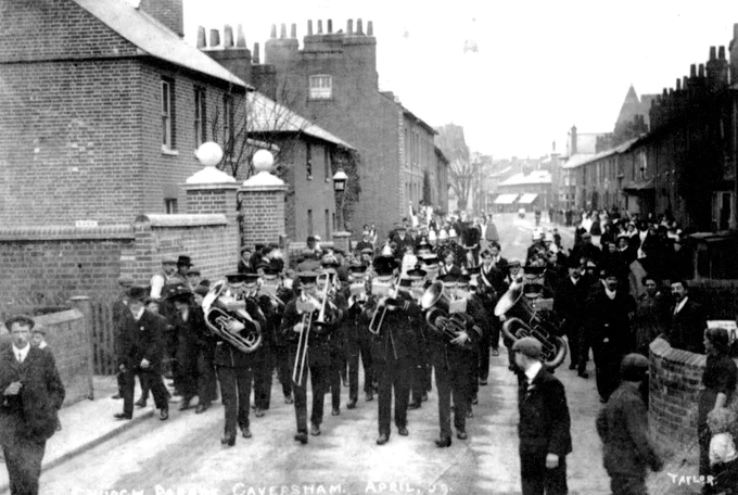 A black and white photo of a brass band marching down a road, with houses either side and some onlookers.