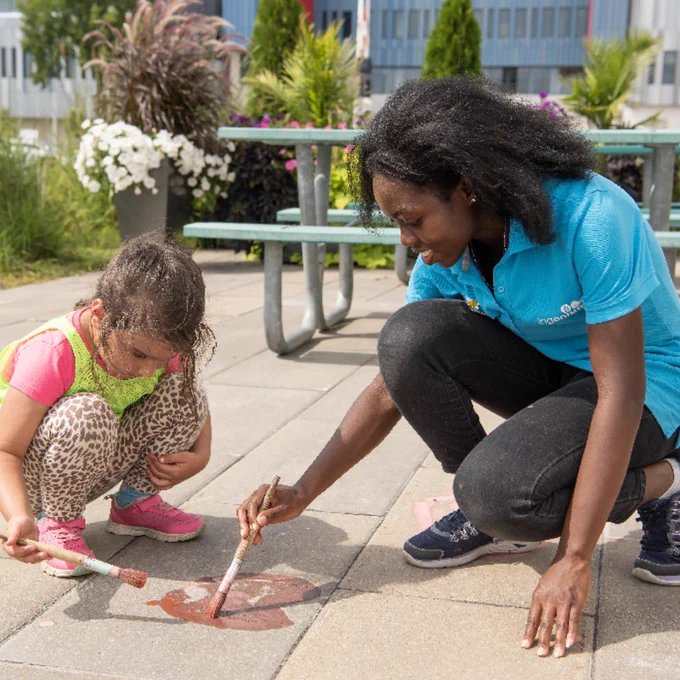 Une femme et une jeune fille sont accroupies sur une surface pavée et dessinent des formes colorées sur le sol à l'aide de pinceaux, en plein air.