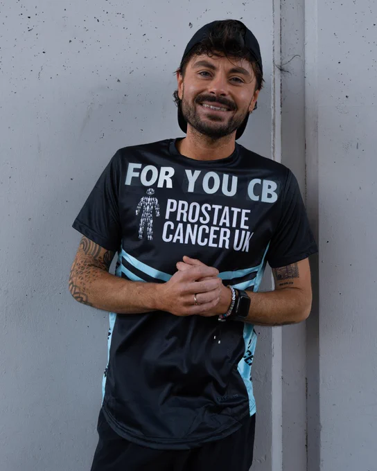 Nick wearing a Prostate Cancer UK running t-shirt, standing against a concrete wall, clasping hands, and smiling.