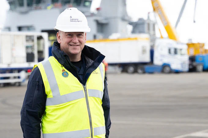 Worker wearing a white hard hat and neon yellow safety vest standing in an industrial port area with trucks and cranes in the background.