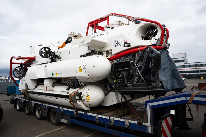 Large white underwater research vehicle with red protective bars loaded on a flatbed trailer at an industrial site.