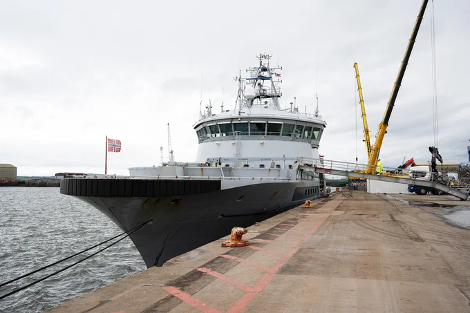 Large naval ship docked at a concrete pier under a cloudy sky with cranes nearby.