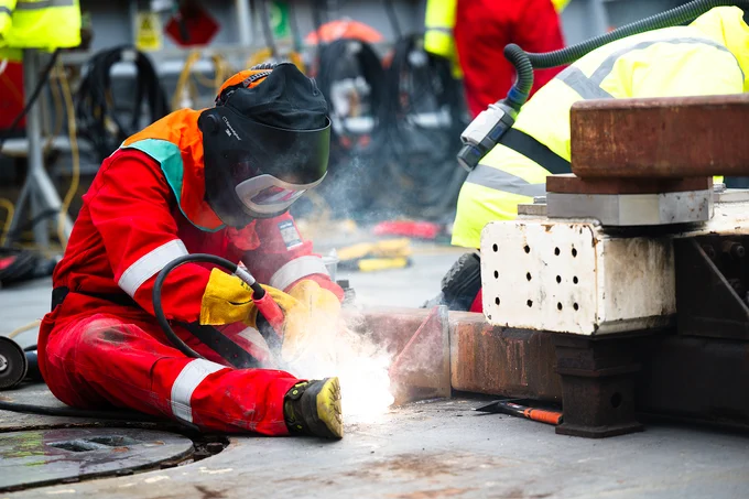 Worker in red protective gear welding metal on an industrial floor with sparks flying.