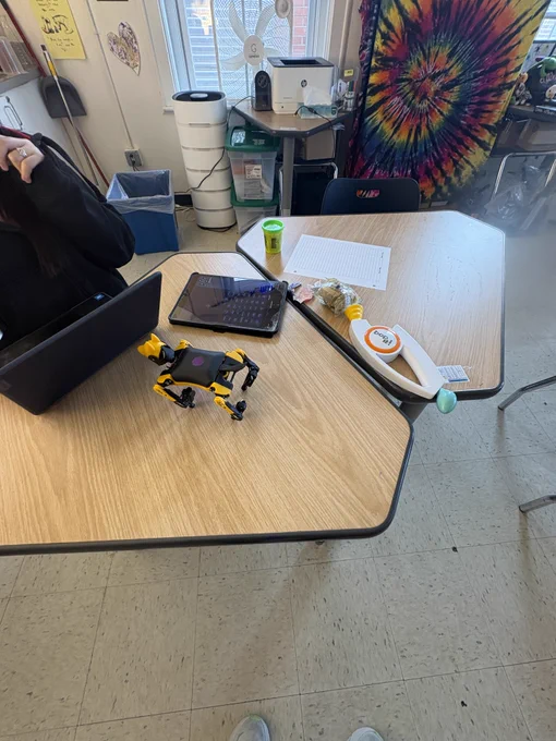A robot dog sits on a classroom desk, being coded by a student on a laptop. 