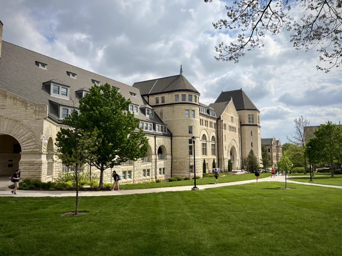 A large, historic stone building with arched windows and a slate roof, part of Kansas State University, set against a partly cloudy sky. The foreground features a well-maintained lawn with scattered trees and a curved walkway along which people are walking.