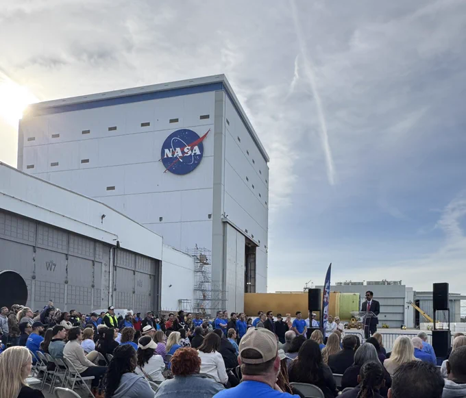 A large group of people watch NASA Associate Administrator Amit Kshatriya talk in a stage during the rollout of the core stage for Artemis 3. The core stage is seen peeking out of the Vertical Assembly Building at NASA Michoud. The sun rises on the left side of the image creating a sun glare over the building.