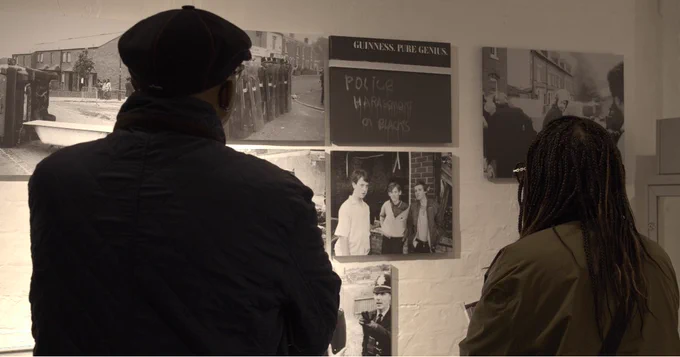 Two people are seen looking towards photographs taken during the Handsworth uprisings