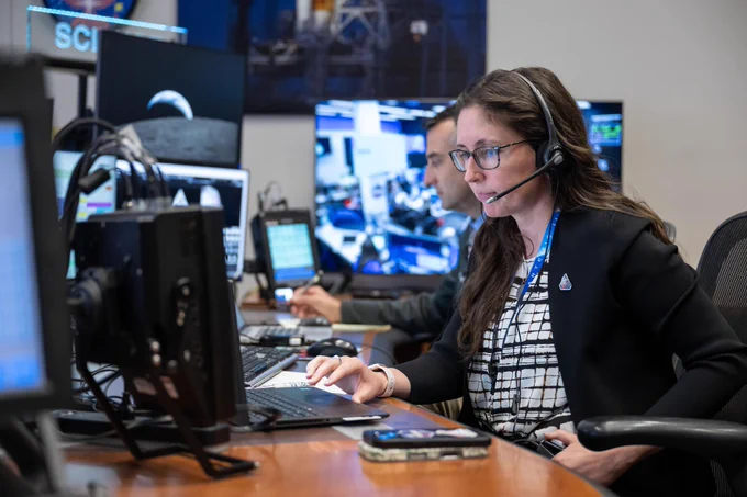 Kelsey Young sits as a desk in mission control, working on a laptop with several monitors behind it. There are many screens visible in the background, showing images of the Moon and data readouts. Kelsey is wearing a headset and has two smartphones stacked on the desk in front of her.  
