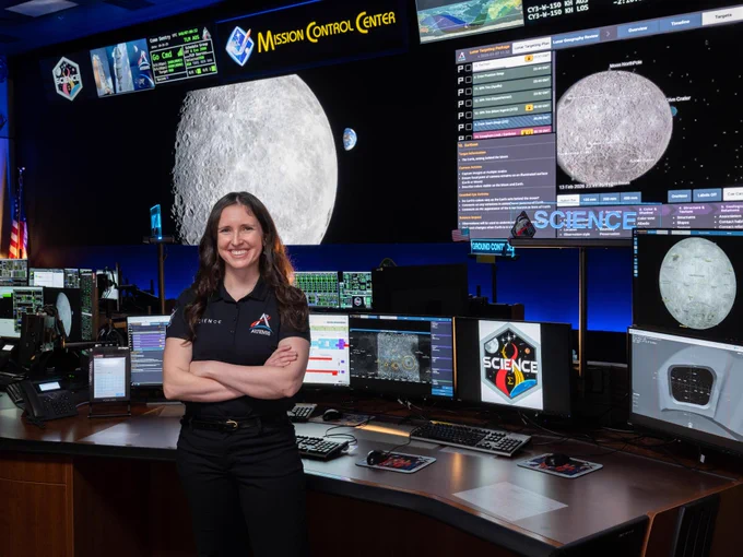 Kelsey Young stands, smiling with her arms crossed in Mission Control. She is wearing an Artemis Science polo, leaning against a curved desk covered in computer monitors. On the wall behind her, massive screens show an image of the Moon along with several different data readouts. A sign along the top of the wall says Mission Control Center. A lit, translucent sign above her monitors says Science.