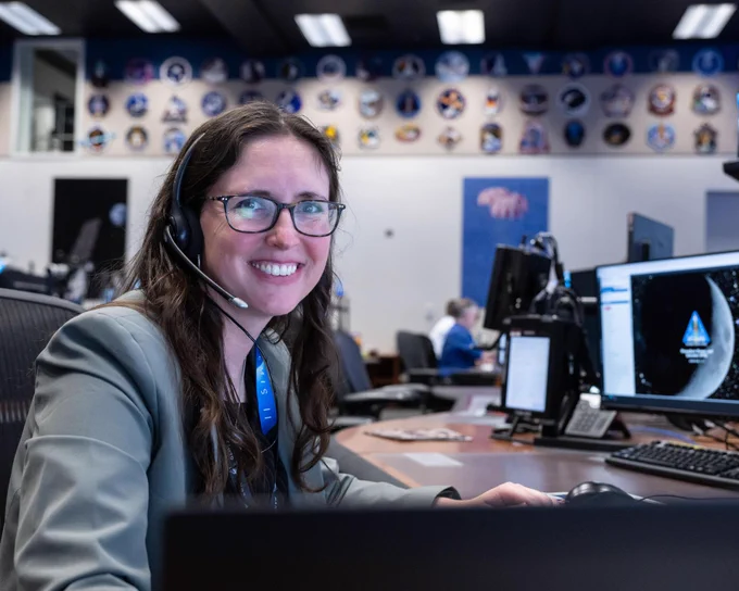 Kelsey Young smiles at the camera while seated at a desk in mission control. The desk is long and stretches along the length of the room, covered with computer monitors. Kelsey is wearing a headset. On the back wall are dozens of mission patches. 