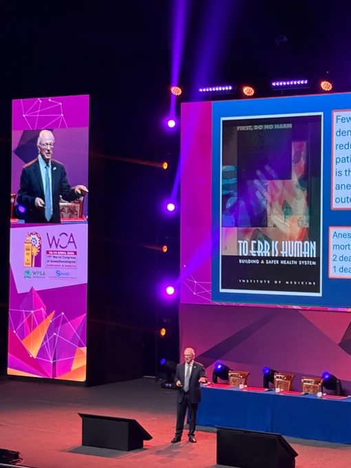 A man in a suit and tie standing on a large stage delivering a lecture  with large banners, lights and projection screen behind him.