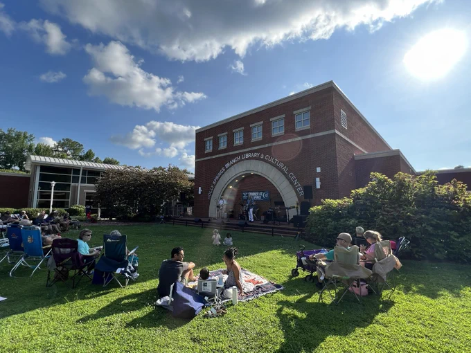 A group of people are enjoying a sunny day on the grass outside the Holly Springs Cultural Center building. The sky is mostly clear with a few clouds.