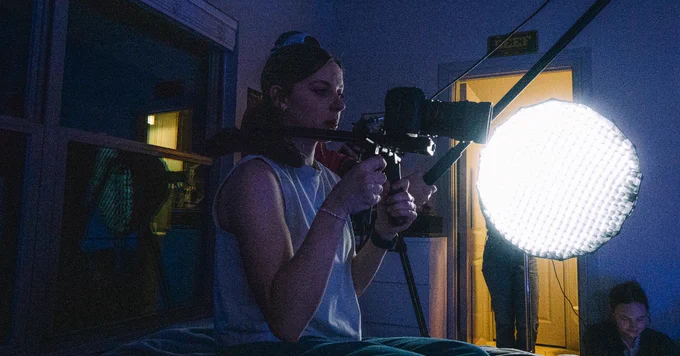 A person operates a camera rig in a dimly lit room, with a bright round light providing illumination. The background includes a door with a small "EXIT" sign above it.