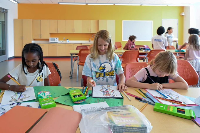 Three children sitting at a table in a classroom, focused on coloring their drawings with colored pencils and markers.