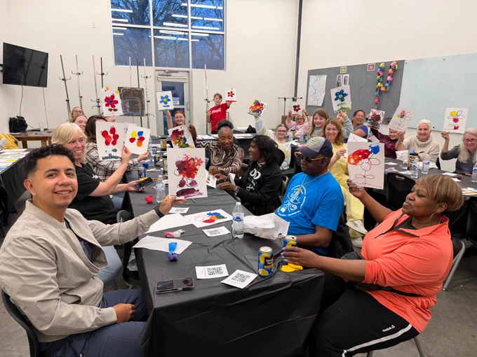 The picture shows a group of adults seated around long tables in a bright room with tall windows. Many people are smiling and holding up their paintings toward the camera. The paintings appear to be colorful, with flower-like shapes and abstract blobs in red, blue, yellow, and purple on white paper. The tables have black tablecloths and are covered with painting supplies and drinks, including several water bottles and a few cans.