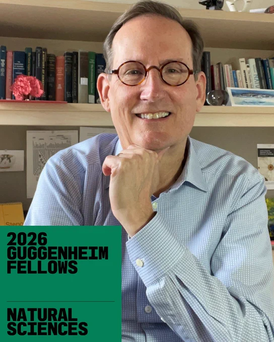A smiling middle-aged man wearing round glasses and a light blue button-down shirt sits in front of a bookshelf filled with books and framed items, resting his chin on his hand. A green graphic overlay in the lower corner reads “2026 Guggenheim Fellows – Natural Sciences.”