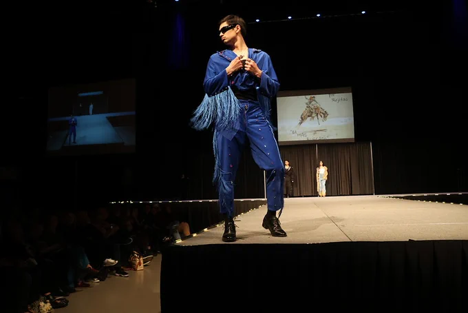 A person stands on a runway wearing sunglasses and a blue outfit. Blue fringe hang long off of each arm.