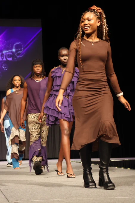 4 people walk up a runway, the lead person wearing a brown dress. Behind her, 3 models wear clothes she designed and made, most featuring purple, gold, or blue.