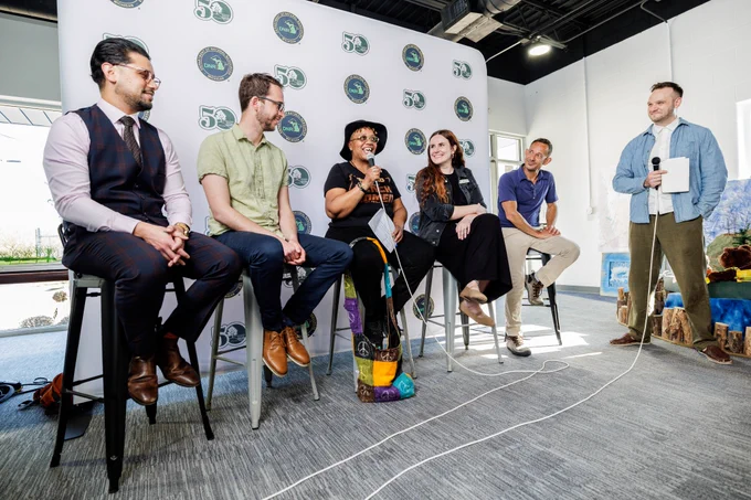 A smiling woman speaks into a microphone as a group seated and standing next to her smile and watch her speak. A logo board is behind the seated group with Michigan Natural Resources Trust Fund 1976-2026 and Michigan Department of Natural Resources logos.