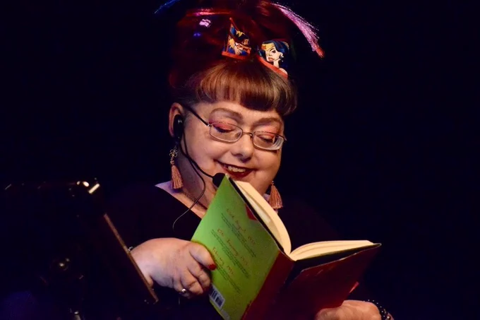Penny, a wheelchair user, is onstage using a headset mic, reading from her memoir First In The World Somewhere, a hardback book with a red front cover and yellow back cover with blurred red and black text. Penny, who wears glasses, is smiling as she looks down. Her red hair is up in a high bun with a wonder woman bow with purple and pink hair extensions. Her face is lit up with a spotlight showing her colourful makeup and her small hands holding open the book. Behind her is a black curtain. 
