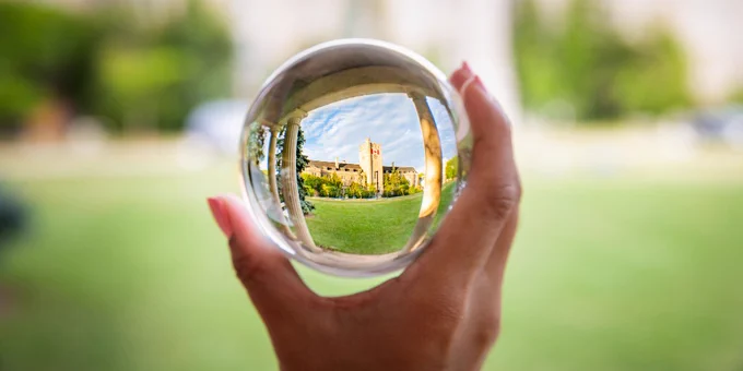 A hand holds a transparent crystal ball in front of Johnston Hall at the University of Guelph; Johnston Hall is out of focus in the background and in focus inside of the crystal ball. 