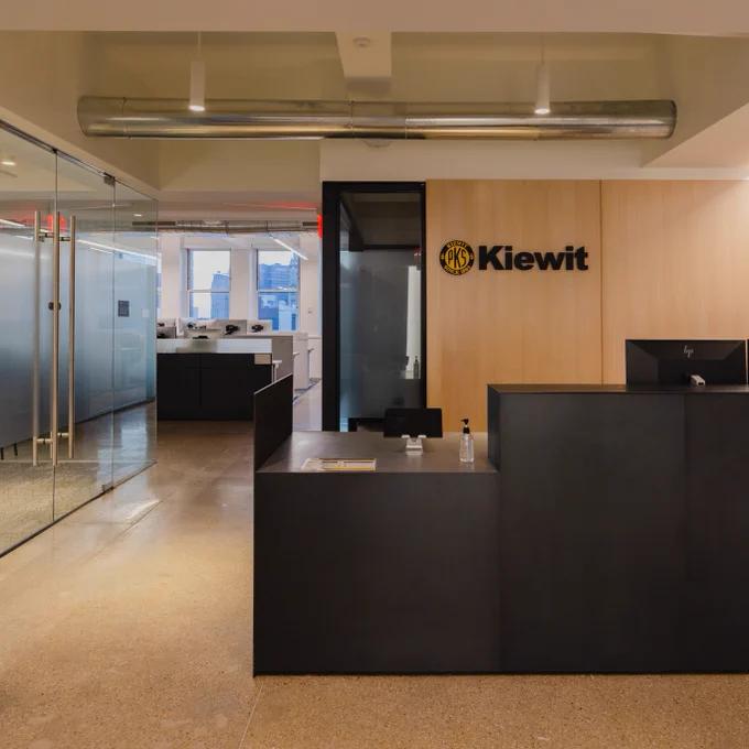 A modern office reception area featuring a large Kiewit logo on a wooden wall. The space includes a sleek black reception desk, a glass-walled meeting room on the left, and minimalist furniture. The area is well-lit with overhead lighting and natural light from windows at the back.