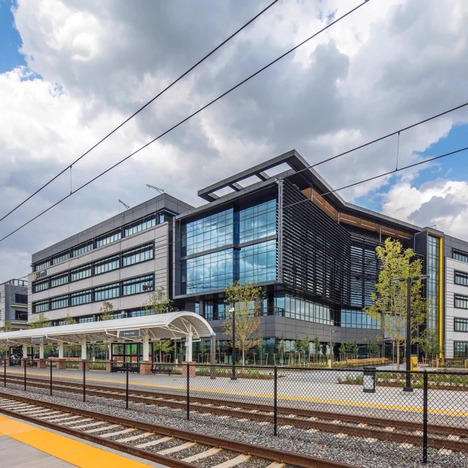 Modern office building surrounded by trees and railway tracks. The front includes a platform and a canopy, with cloudy skies overhead.