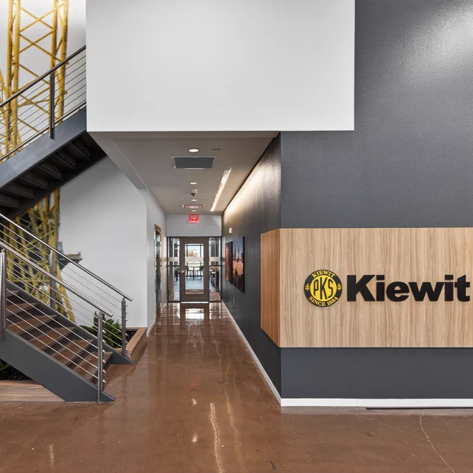 A modern interior hallway with polished floors, featuring a staircase with metal railings on the left. The wall on the right displays the Kiewit logo on a wooden panel. The end of the hallway is brightly lit, and plants are arranged at the base of the staircase.