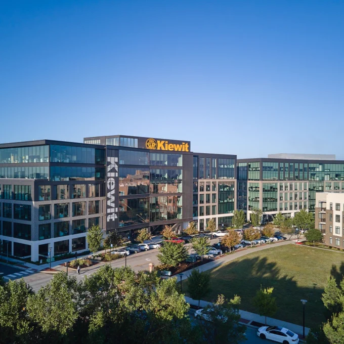 A modern office building complex with large glass windows, prominently featuring the Kiewit logo on the top. The complex is surrounded by trees and a grassy area, with several parked cars visible near the entrance.